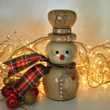 A wooden snowman figurine with a plaid red, green, and black ribbon scarf, decorated with small rhinestones and a red nose, set against a warm background with glowing fairy lights and holiday ornaments.