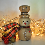 A wooden snowman figurine decorated with a red and green plaid ribbon, small rhinestones, a red nose, and a black painted face, styled with warm white fairy lights and holiday ornaments on a light background.