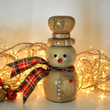 A wooden snowman figurine with a red plaid scarf and a red nose, decorated with small rhinestones, set against a warm background with glowing fairy lights.
