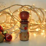 A small wooden snowman figurine with a red hat, red nose, and silver scarf, styled with warm white fairy lights and red ornaments on a light background.