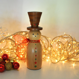 A wooden snowman figurine with a dark brown top hat, a red ribbon bow, and red and green decorative buttons, styled with warm white fairy lights and holiday ornaments on a light background.