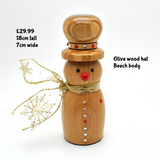 A wooden snowman figurine with an olive wood hat, a beech body, a gold glitter scarf, and small red and silver decorative accents, shown on a white background with text overlays detailing its size and materials.