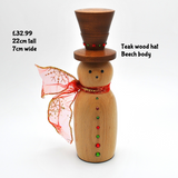 A handmade wooden snowman figurine made of teak wood with a brown hat, a red ribbon scarf, and small red and green decorative gems. Text overlays specify the dimensions and materials.