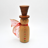 A wooden snowman decoration with a dark brown top hat, a light natural wood body, red and green decorative buttons, and a red glittery scarf, styled with warm fairy lights, gold beads, and holiday ornaments.
