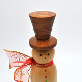 A wooden snowman decoration with a dark brown top hat, a light natural wood body, red and green decorative buttons, and a red glittery scarf, styled with warm fairy lights, gold beads, and holiday ornaments.