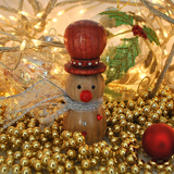 A wooden snowman ornament with a red hat, red nose, and silver scarf, surrounded by gold beads, fairy lights, and Christmas decorations.