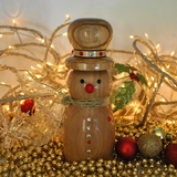 A wooden snowman ornament or decorative figure with a top hat, black eyes, a red nose, gold scarf, and small red and white details, styled with warm string lights, gold beads, and Christmas ornaments.