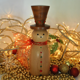 A wooden snowman decoration with a dark brown top hat, a light natural wood body, red and green decorative buttons, and a red glittery scarf, styled with warm fairy lights, gold beads, and holiday ornaments.